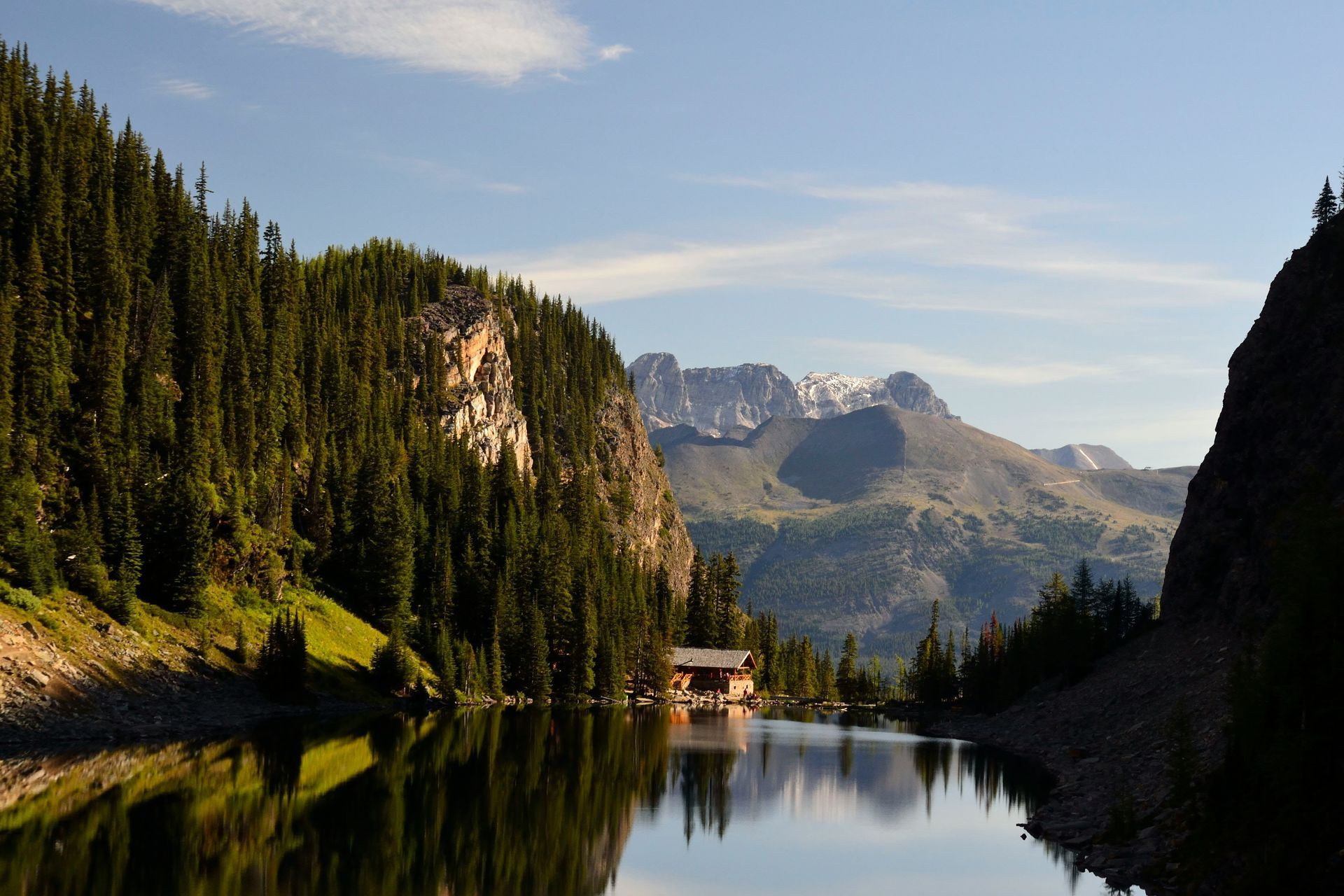Mountain lake framed by pine forest and cliffs, with a small cabin reflecting in calm water at sunset