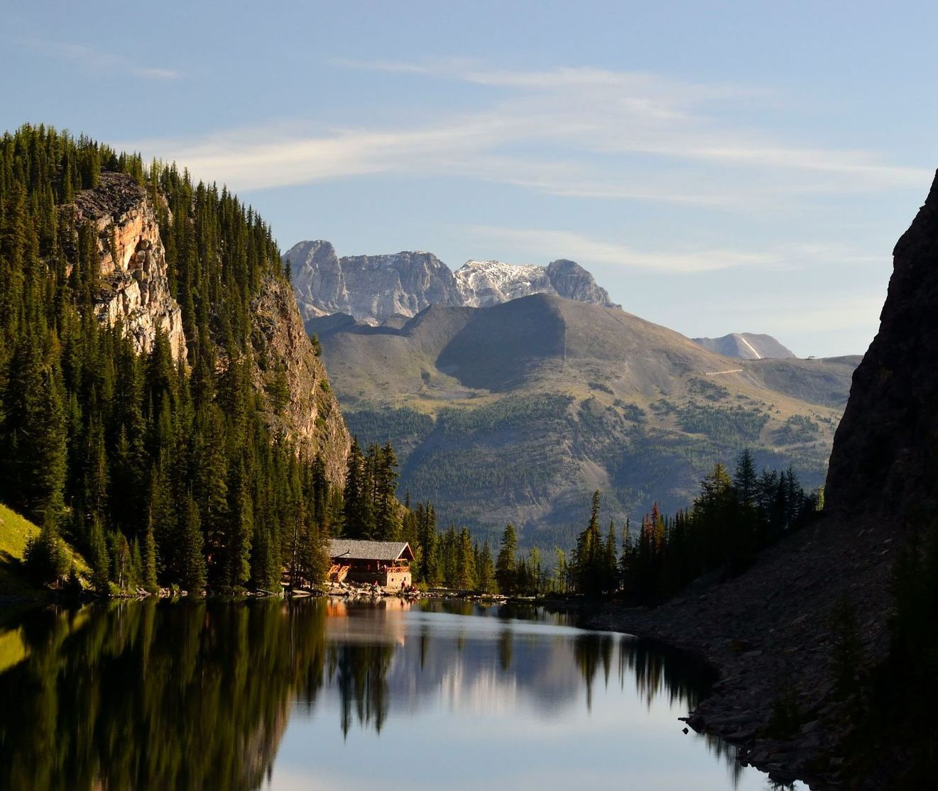 Mountain lake with evergreen forest, rocky cliffs, and a small cabin reflected in calm water