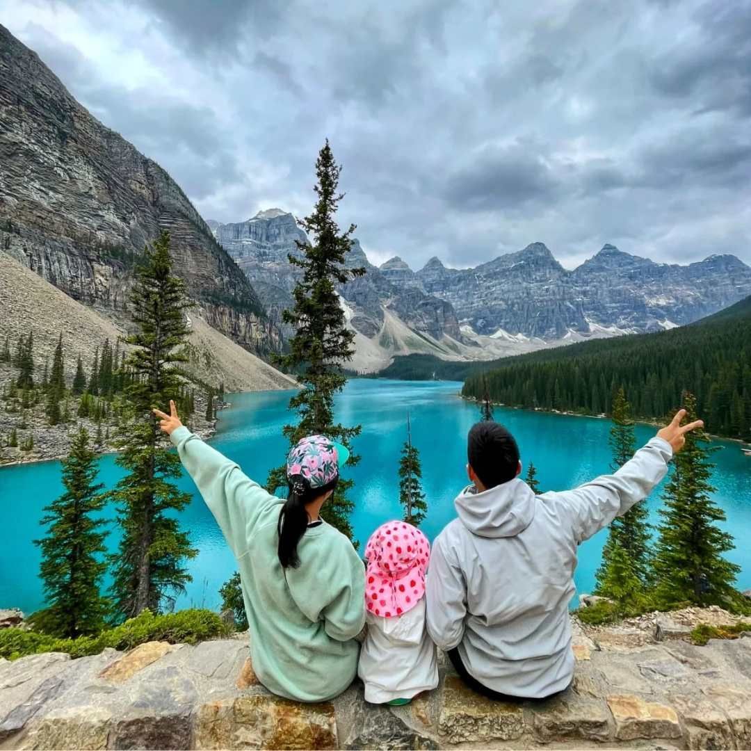 Three people sit by a turquoise alpine lake, arms raised, surrounded by mountains and pine trees.