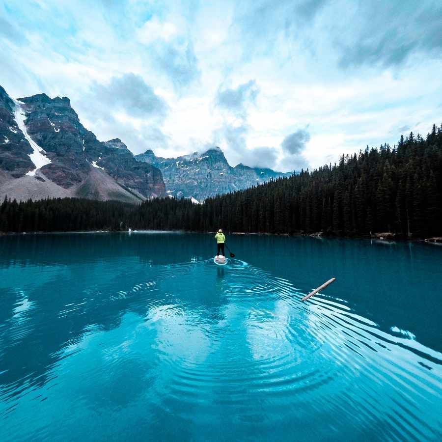 Person paddleboarding on a turquoise mountain lake with snowy peaks and pine forest in the background