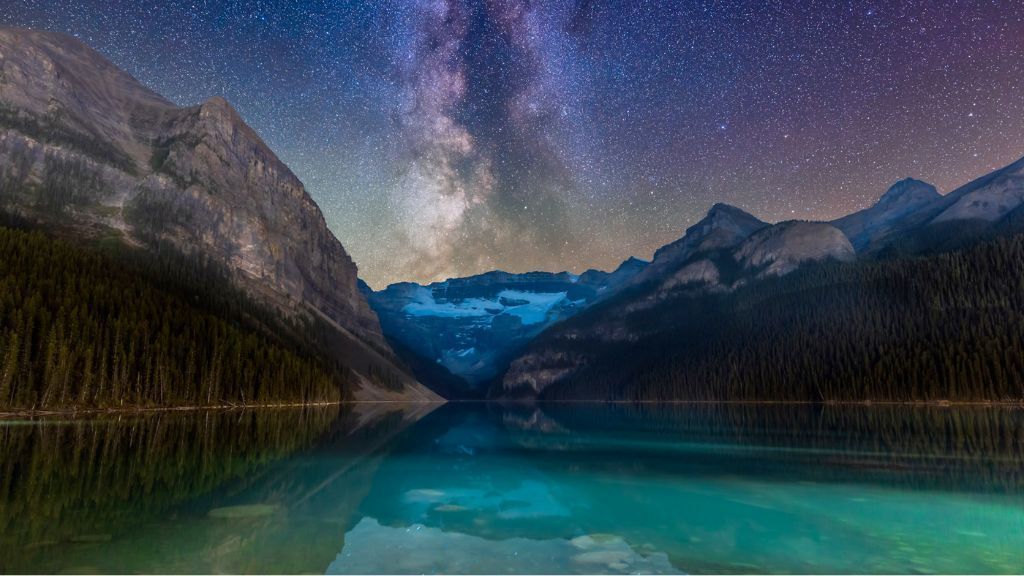 Starry night sky over a mountain lake with mirrored peaks and a glowing Milky Way