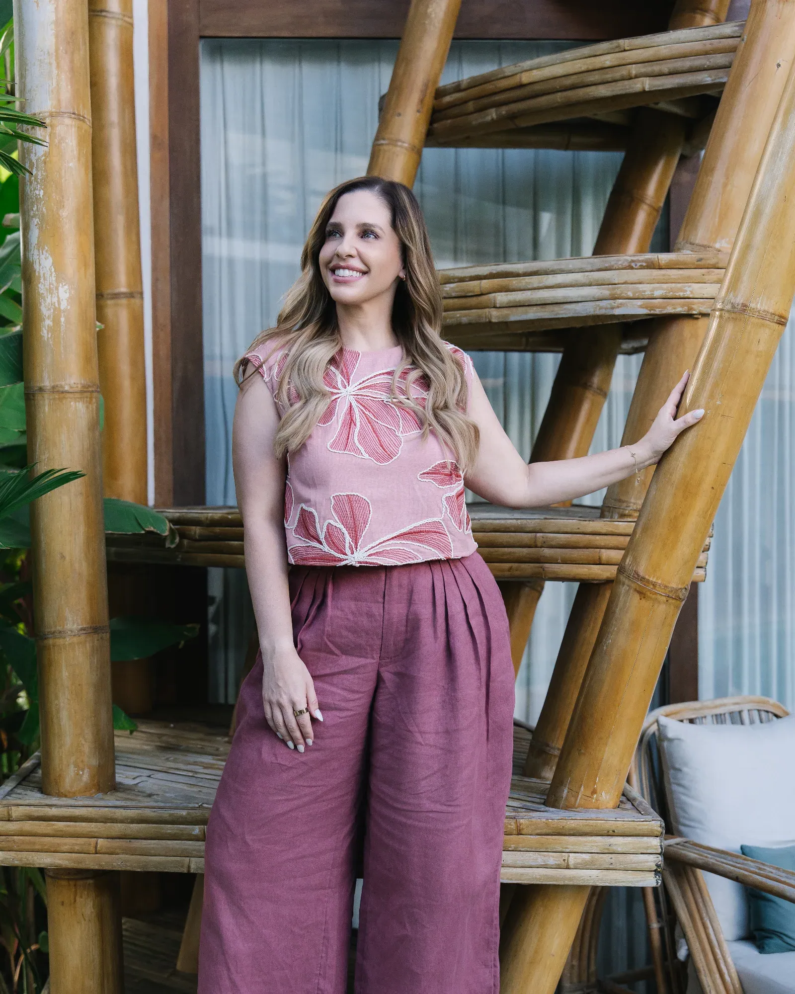 Woman with blonde hair smiles, wearing white shirt and jeans, sitting in front of abstract pink and red background.