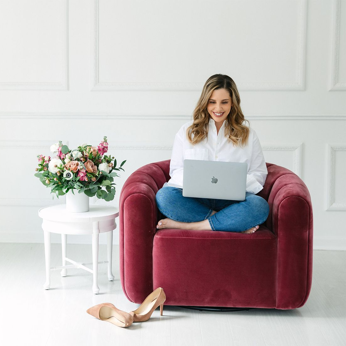 Woman sitting cross-legged in a red chair, working on a laptop. White shirt, blue jeans, shoes on the floor.