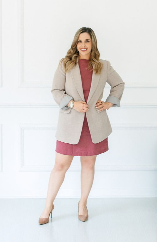 Woman in a red dress and tan blazer smiles, posing against a white wall.