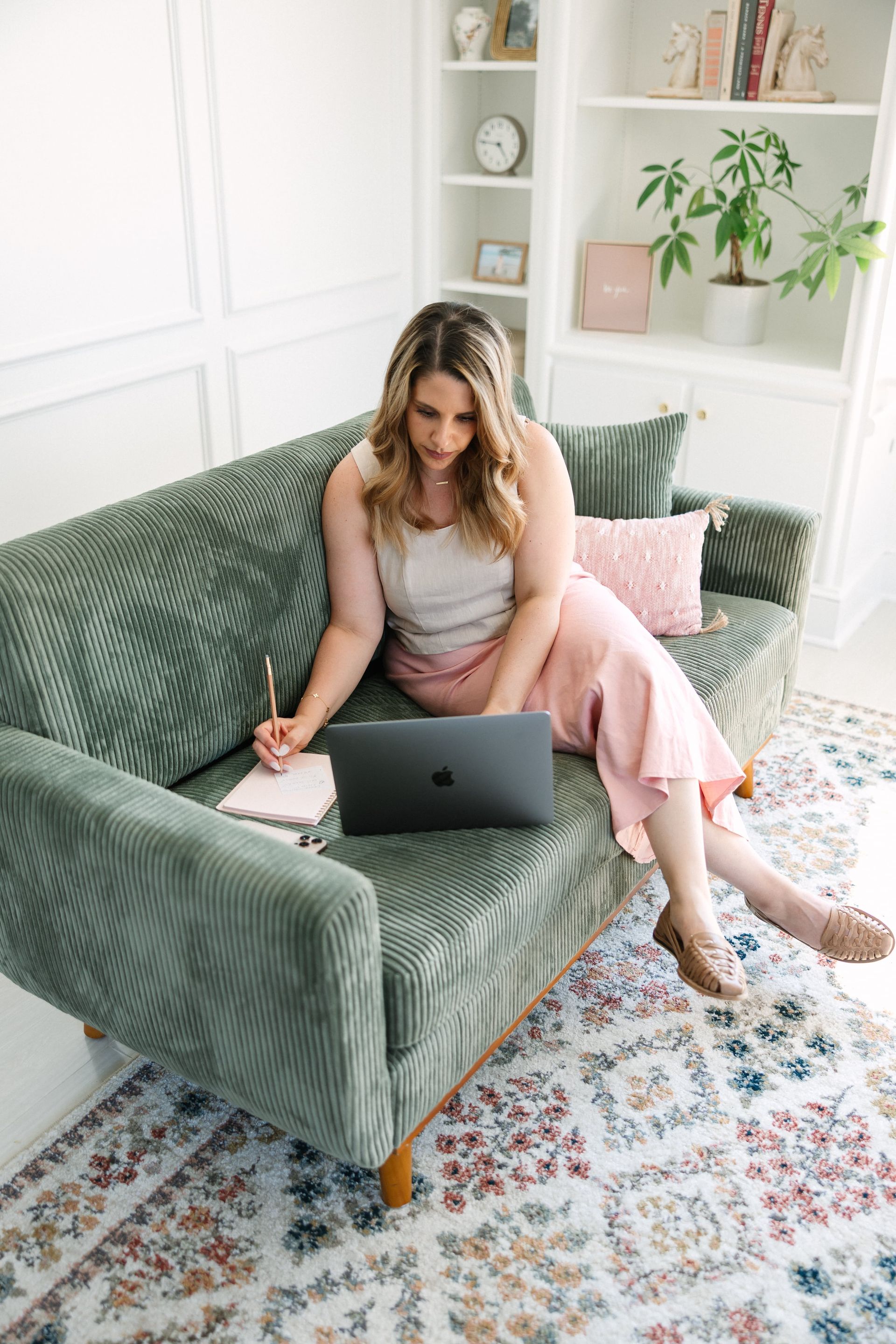 Woman working on laptop, taking notes on a green couch.  Light-filled room.