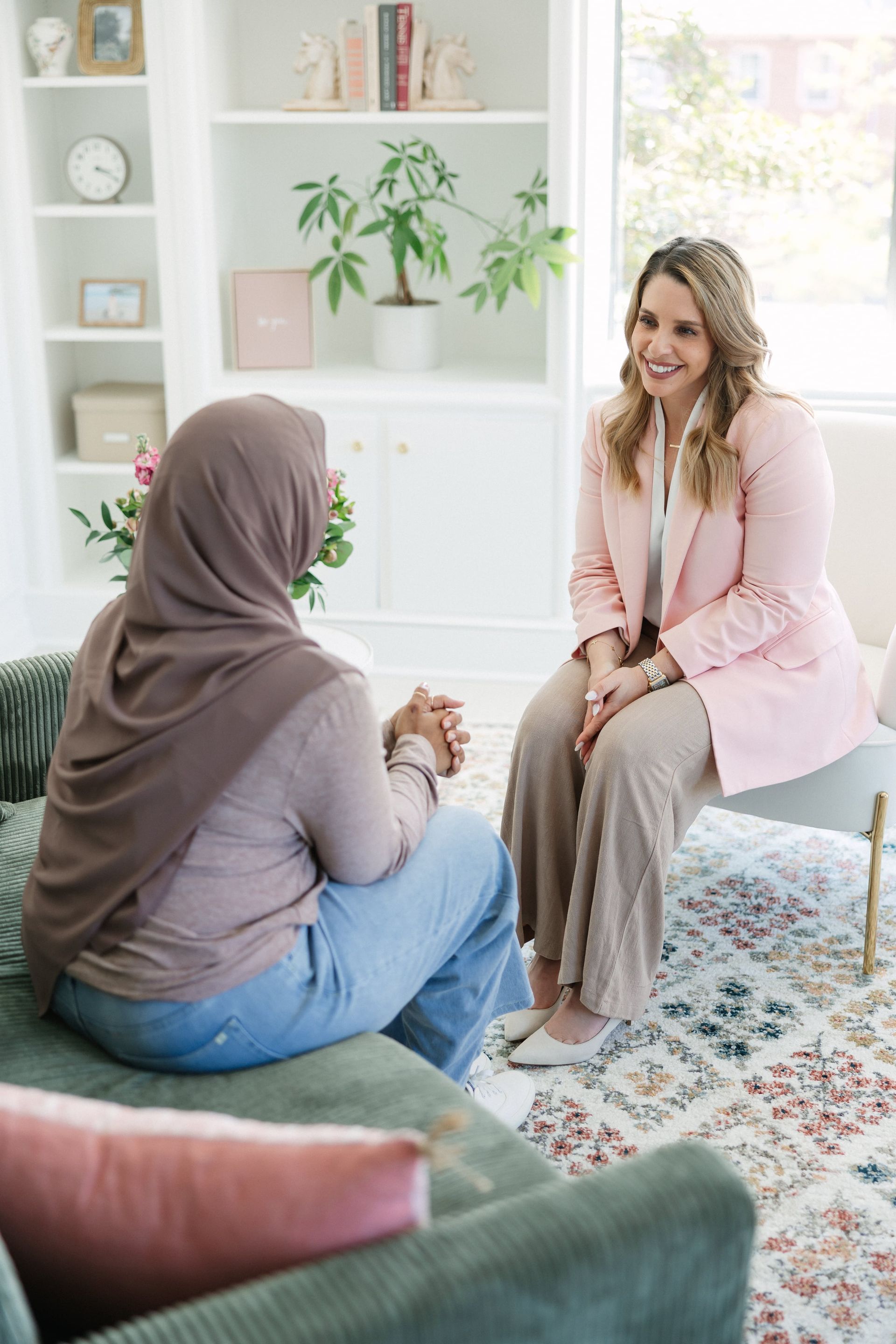 Woman in hijab on sofa, facing smiling woman in pink blazer and pants in a bright room.