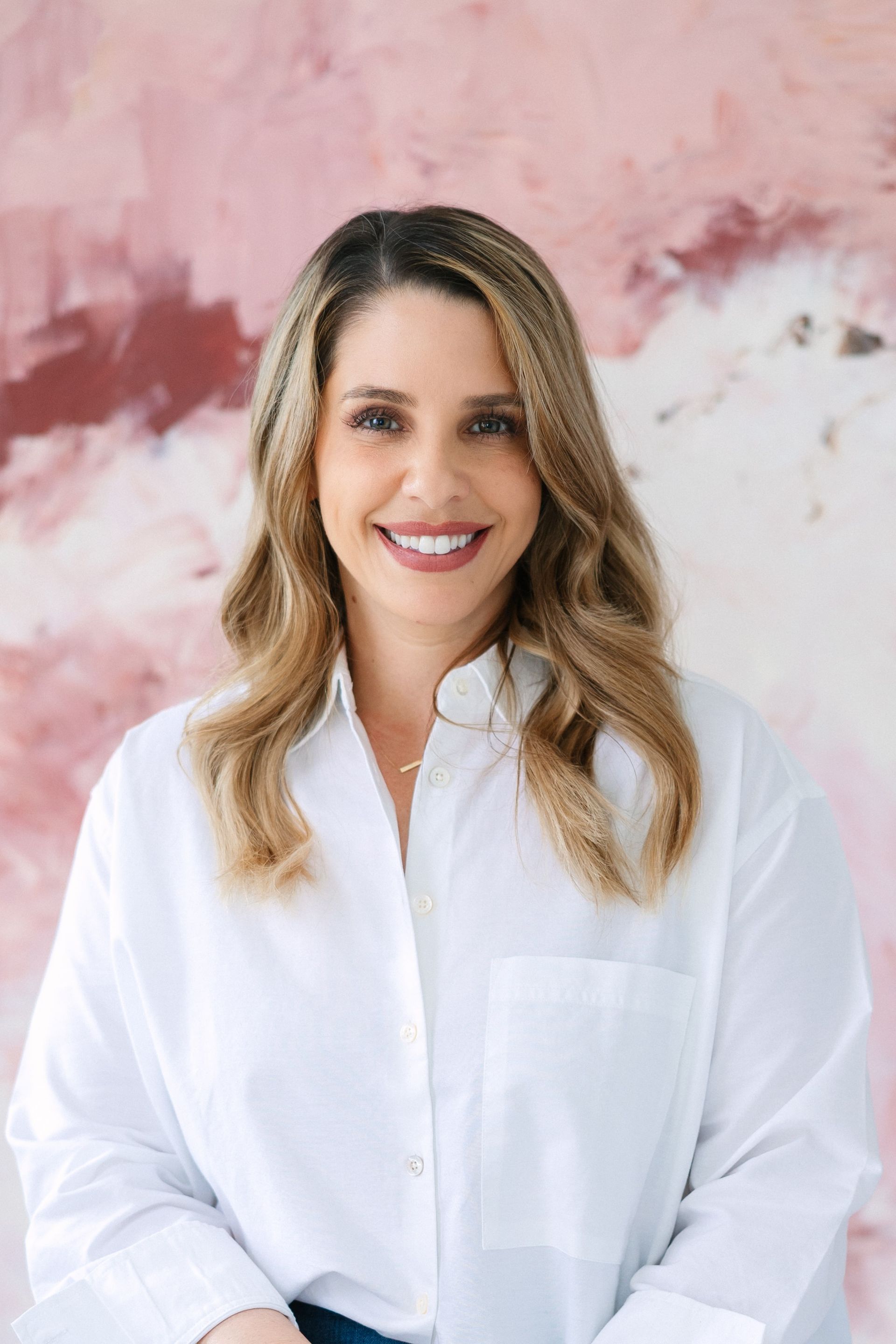 Woman smiling, wearing a white button-down shirt, in front of a pink abstract painting.