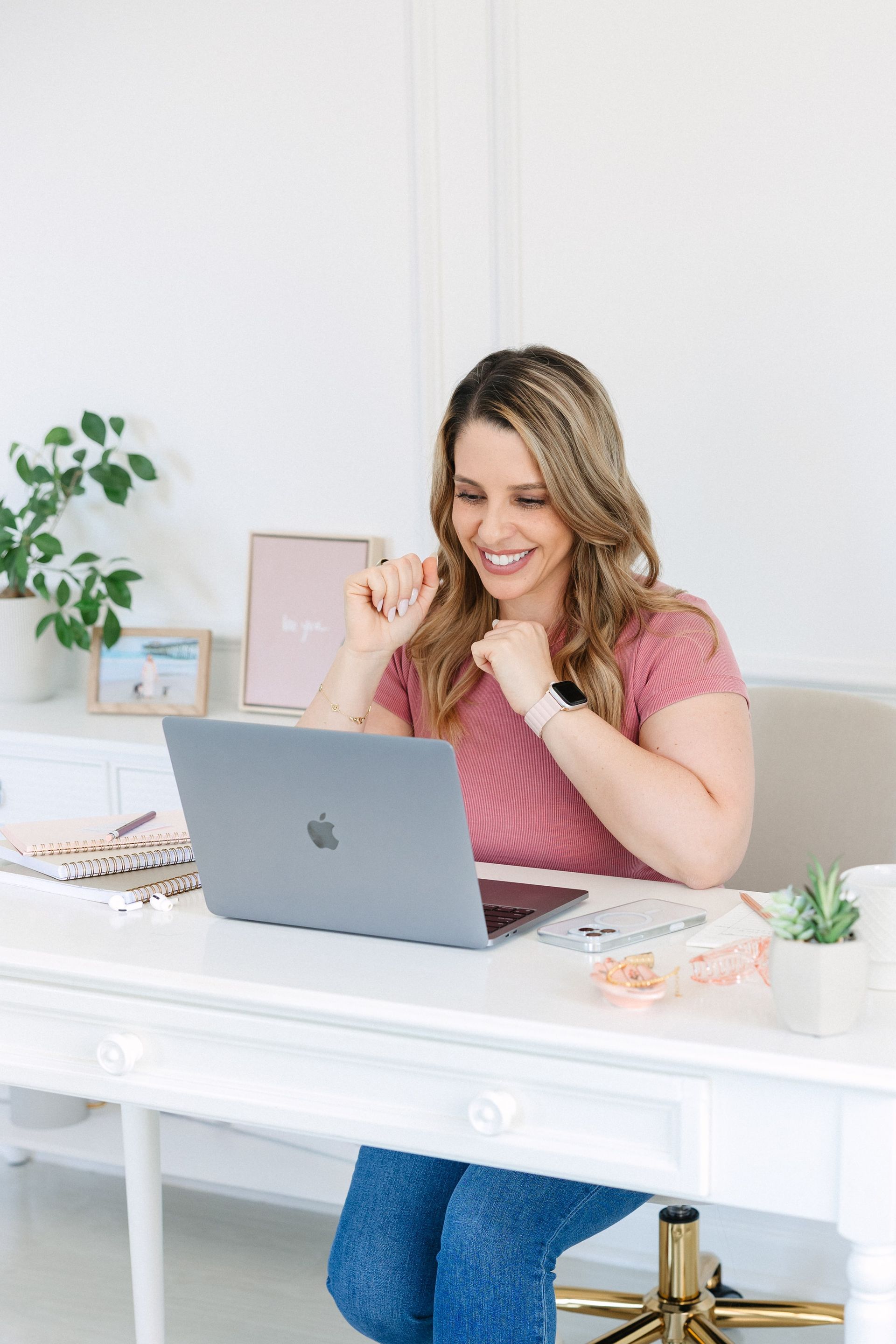 Woman smiles while looking at laptop, sitting at white desk.