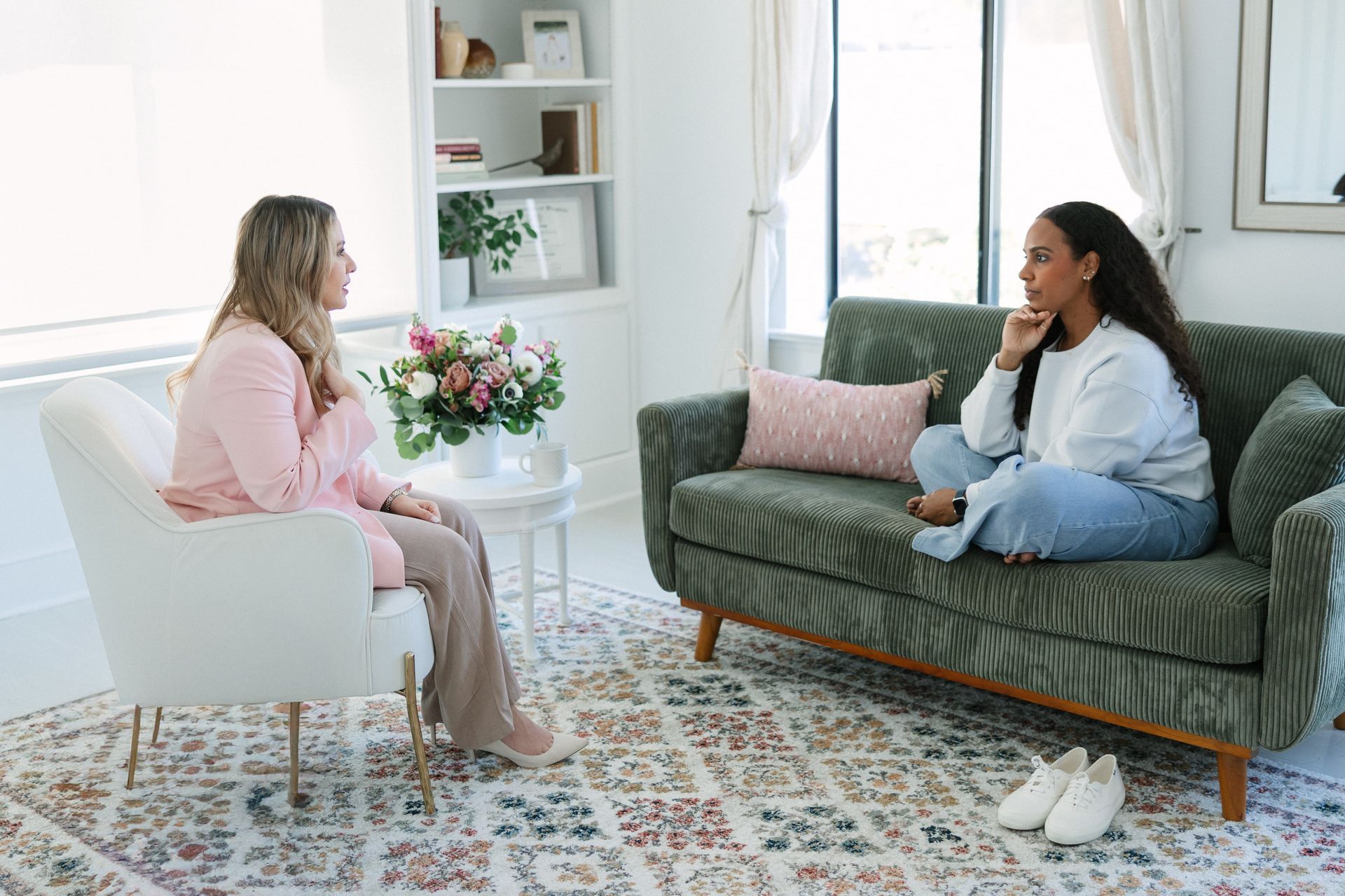 Woman in pink jacket seated, talking to another woman on a couch. They are indoors in a bright room.