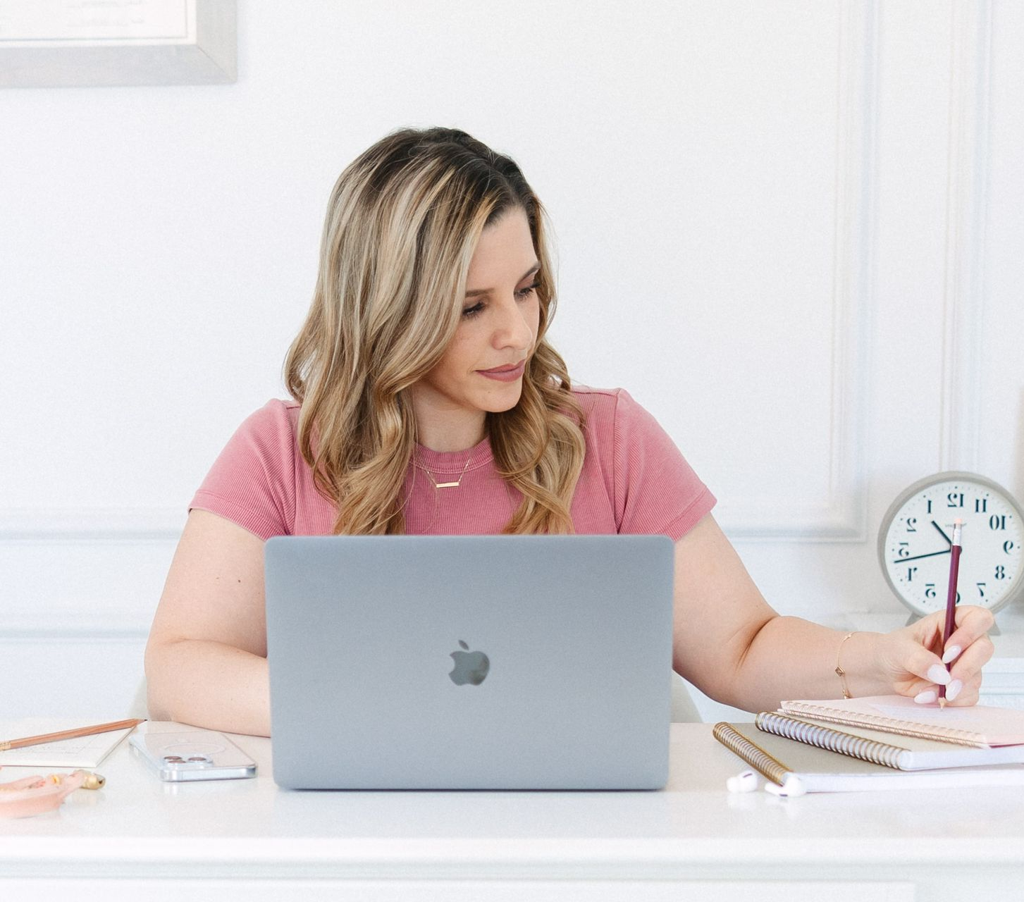 Woman at a desk with laptop, writing in notebook. Light pink shirt, neutral tones.