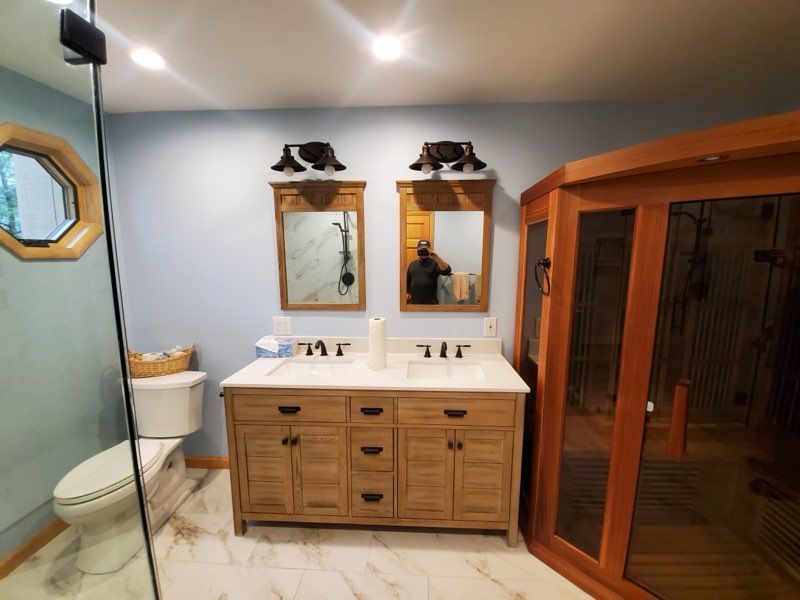 Bathroom with double vanity, two mirrors, sauna, and a toilet. Light blue walls, wood cabinetry.