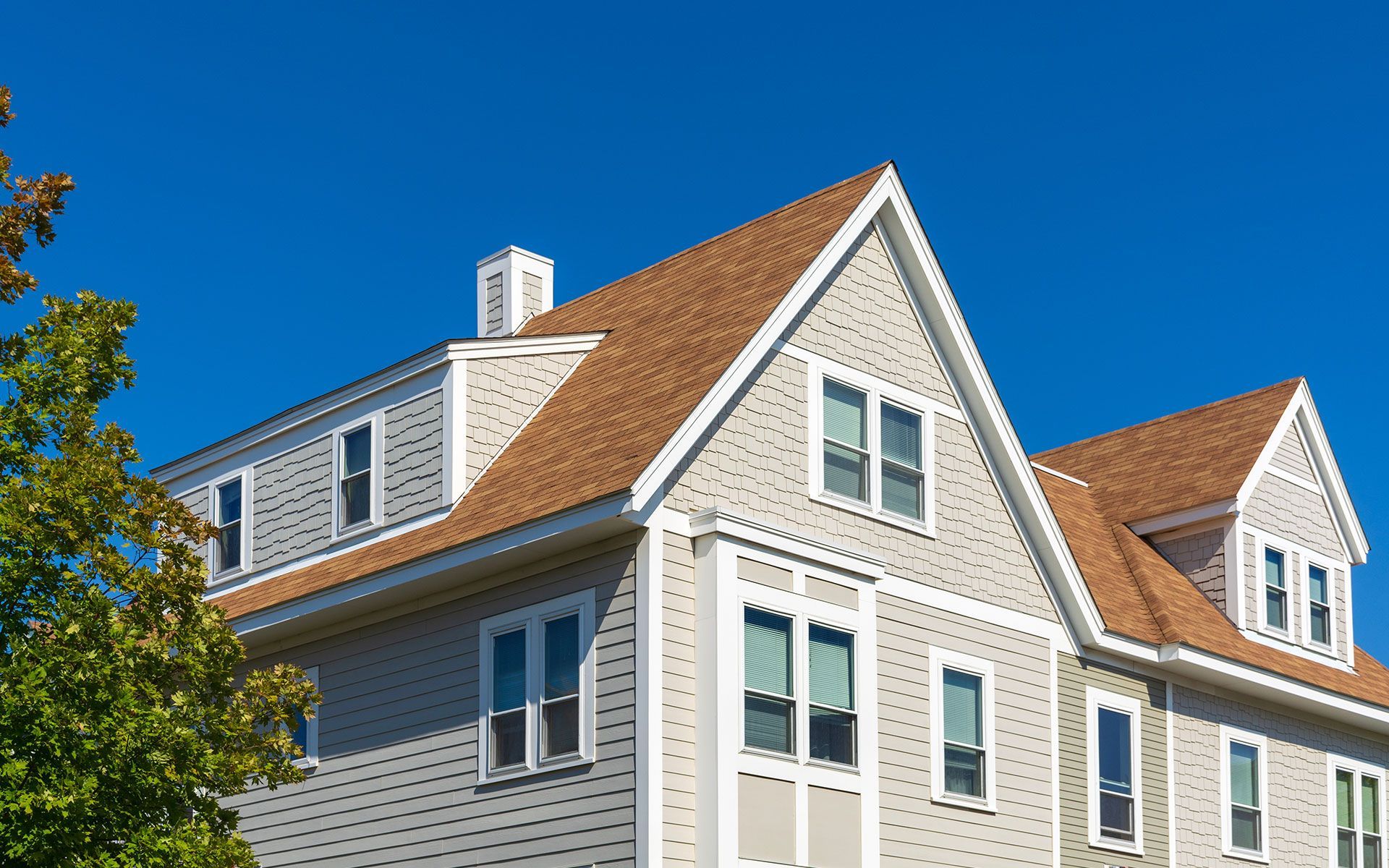 Two-story house with light grey siding, brown roof, and small dormers against a bright blue sky.