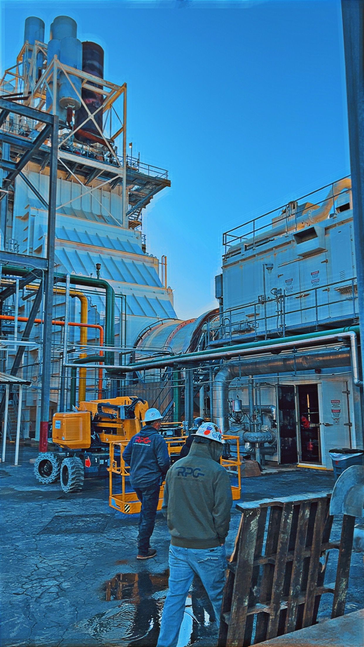 Industrial plant with two workers in hard hats beside blue steel structures and pipes outdoors