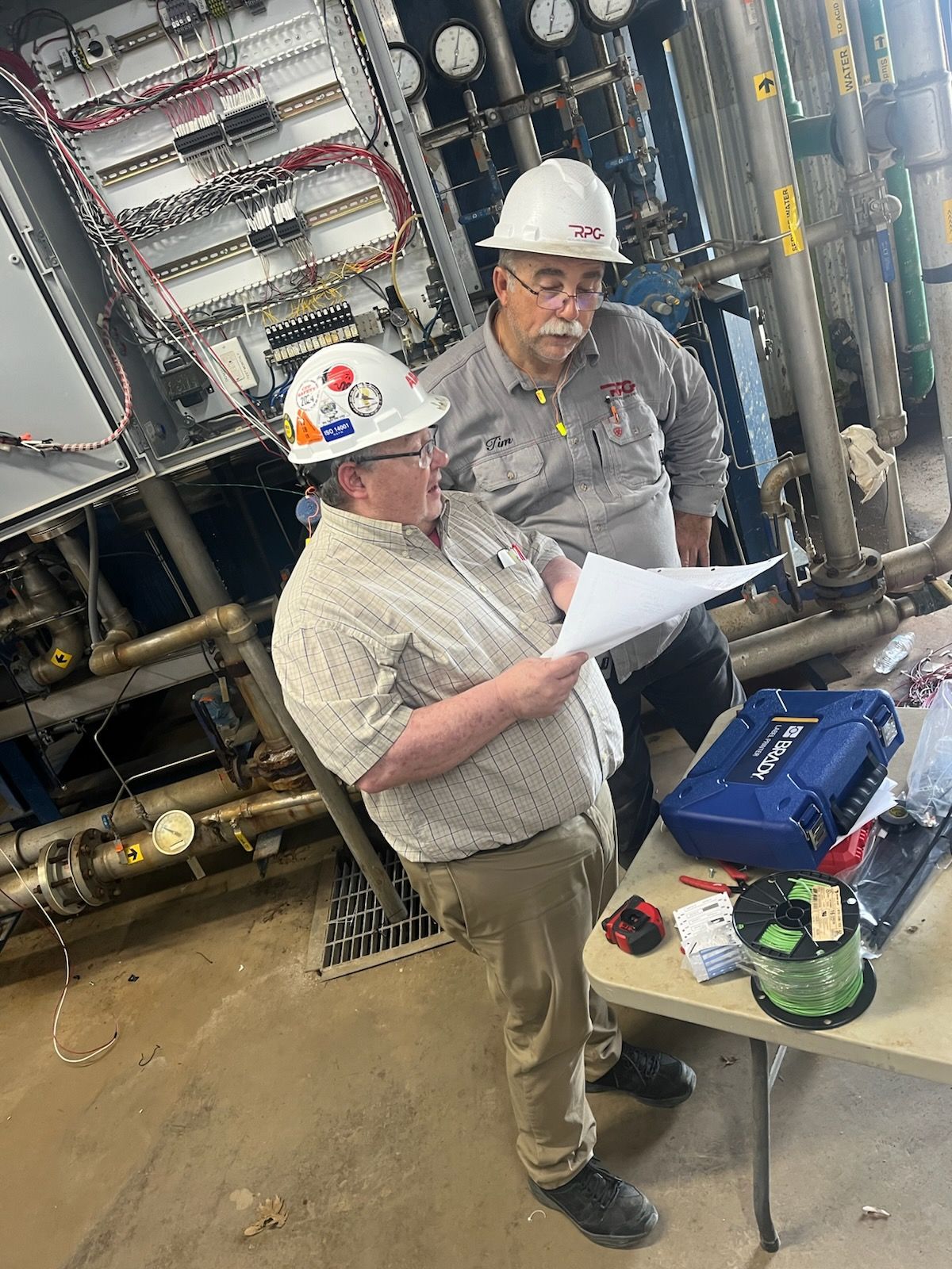 Two workers in hard hats reviewing paperwork beside industrial equipment and a table with tools.