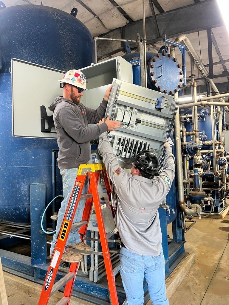 Two workers on ladders installing equipment in an industrial plant beside large blue tanks.