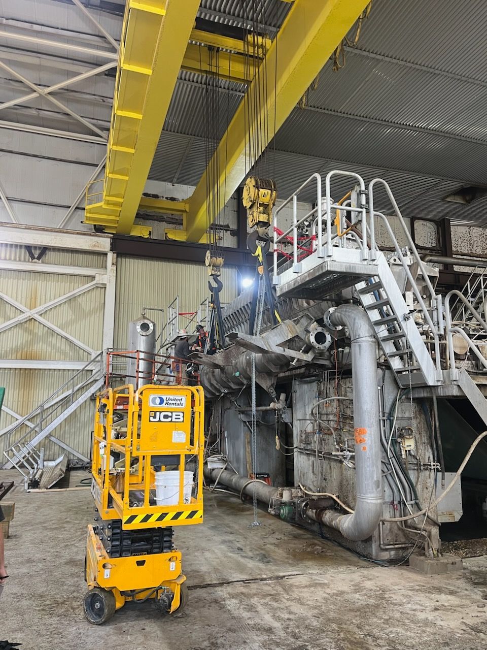 Yellow lift in industrial hangar beside large machinery and worker on elevated platform.
