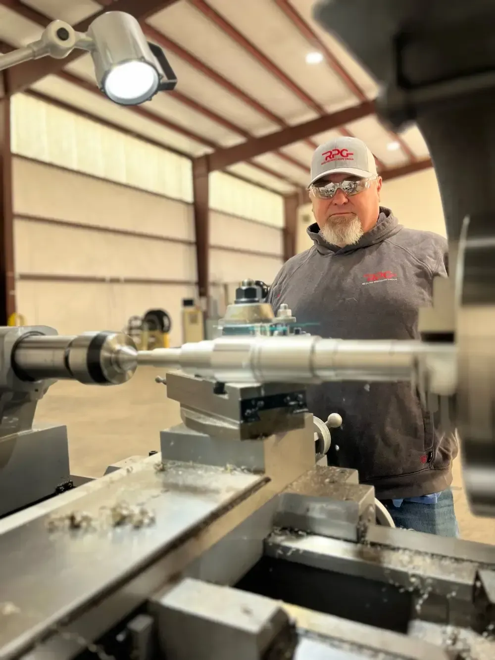 Man operating a metal lathe in a workshop, with a long silver workpiece and tools in the foreground.