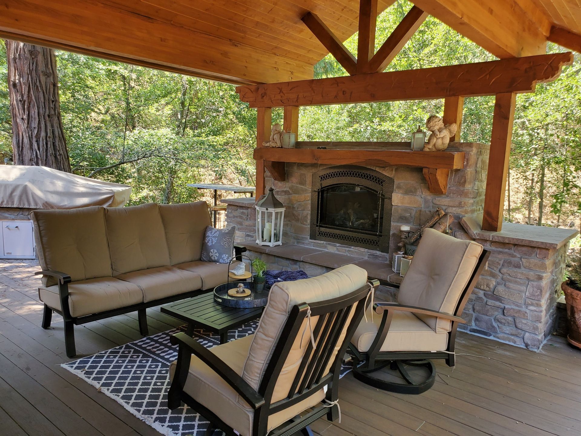 Covered outdoor patio with cushioned chairs, glass table, and stone fireplace under a wooden roof