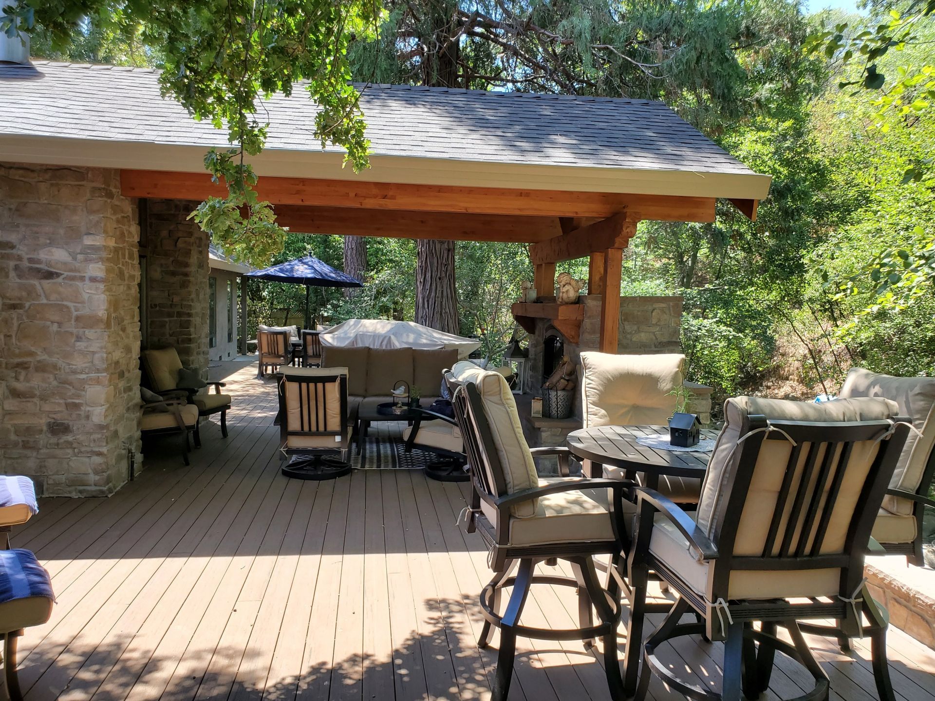Covered backyard patio with outdoor seating, wooden deck, stone columns, and trees in the background