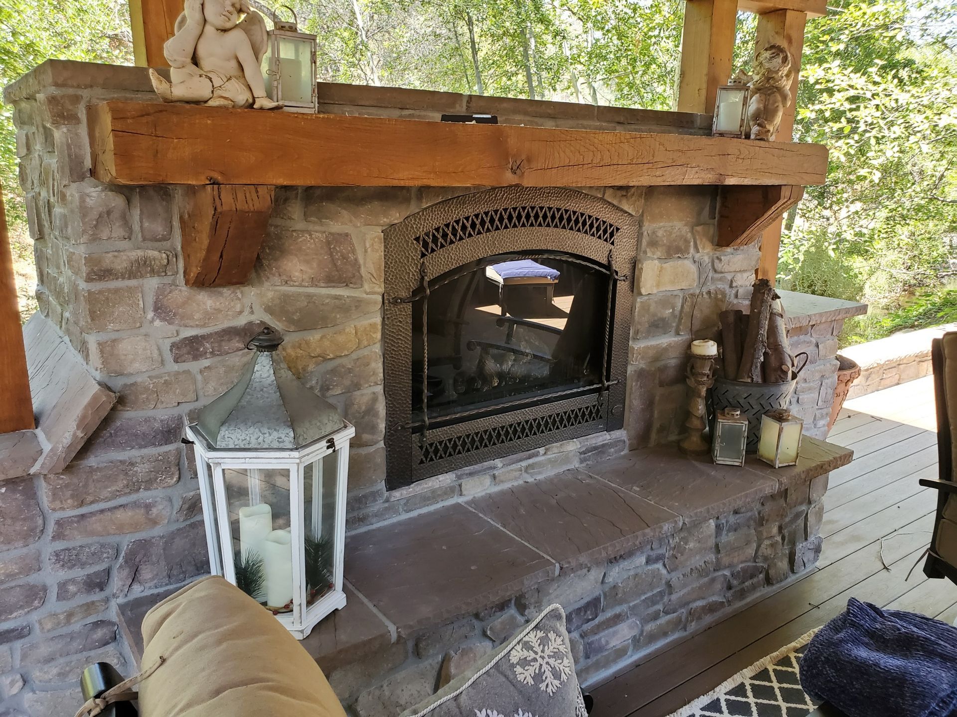 Stone outdoor fireplace with wooden mantel, lanterns, and a glass fire screen on a porch.