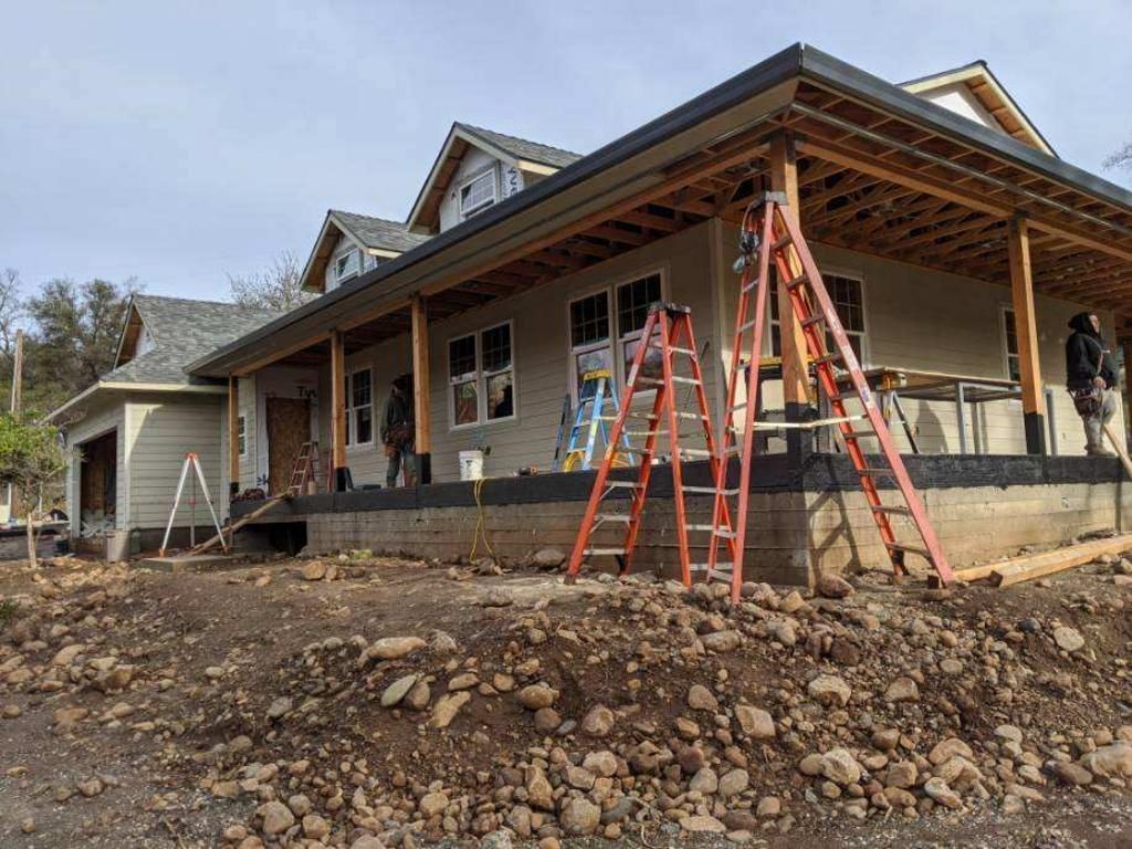 House under construction with ladders on a dirt lot and workers on the porch
