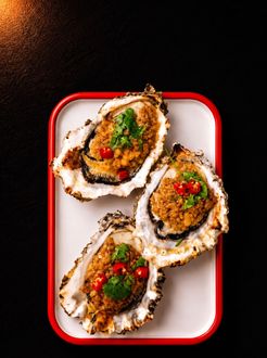 Three baked oysters on a white tray with red rim, topped with herbs and diced red peppers