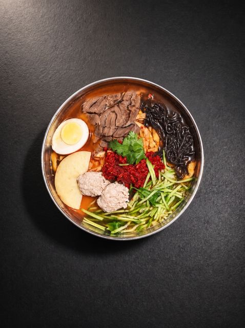 Ramen bowl with sliced egg, pork, seaweed, corn, greens, and red garnish on a dark table
