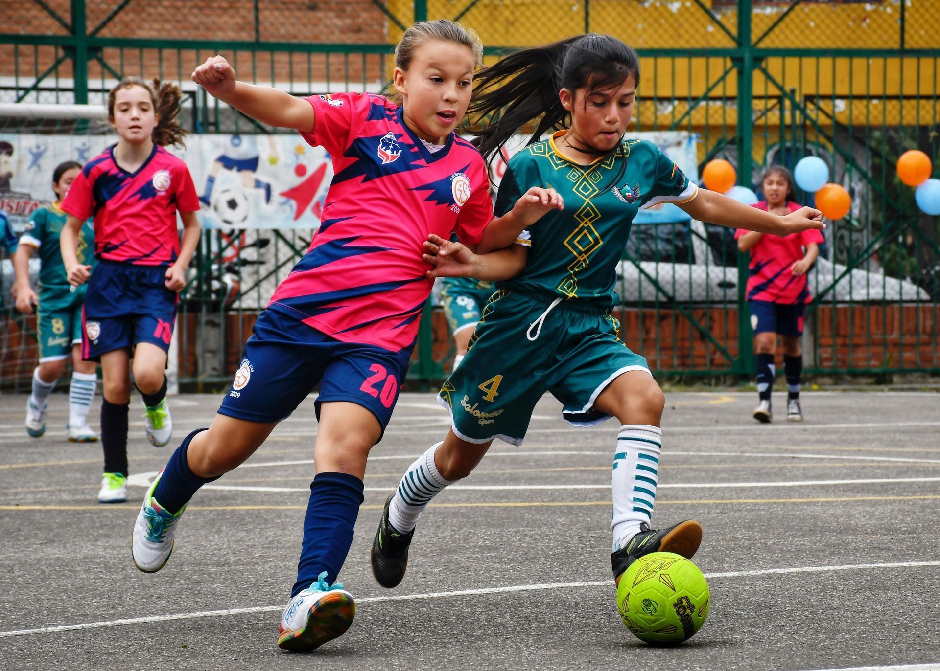 Two girls in soccer uniforms compete for the ball on a blacktop, others watch.