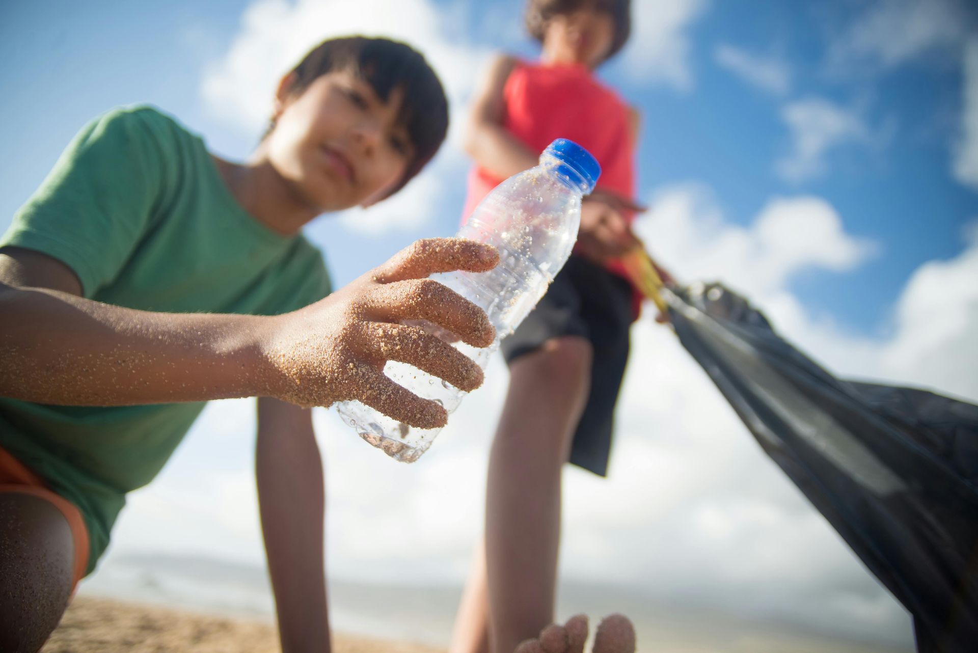 Two children picking up trash on a beach; one holding a water bottle, the other with a trash bag.