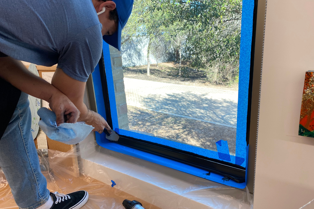 A man painting a window with blue tape, preparing it for security film installation.