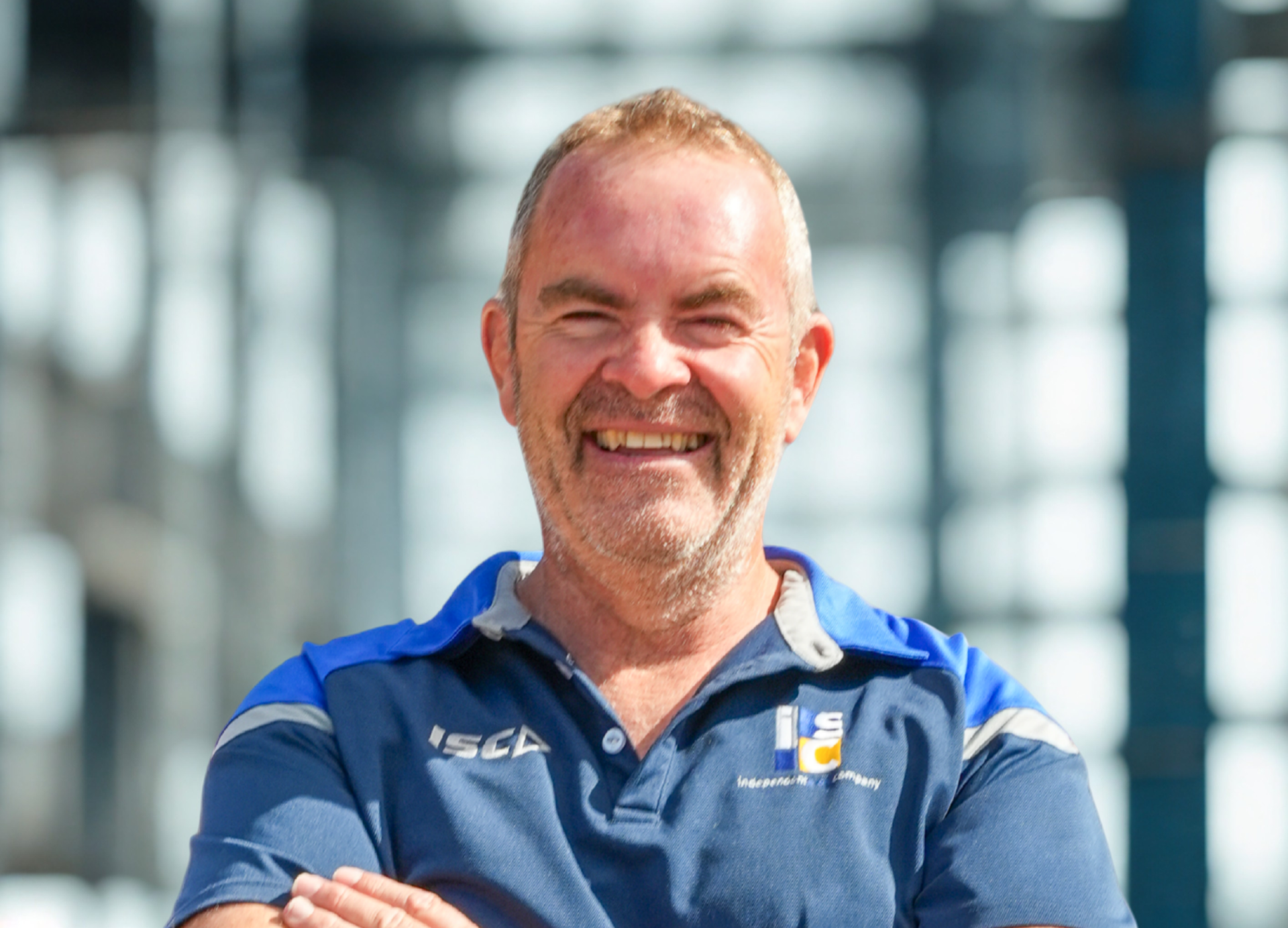 A smiling person with short hair, wearing a blue polo shirt, stands with arms crossed against a blurred industrial backdrop.