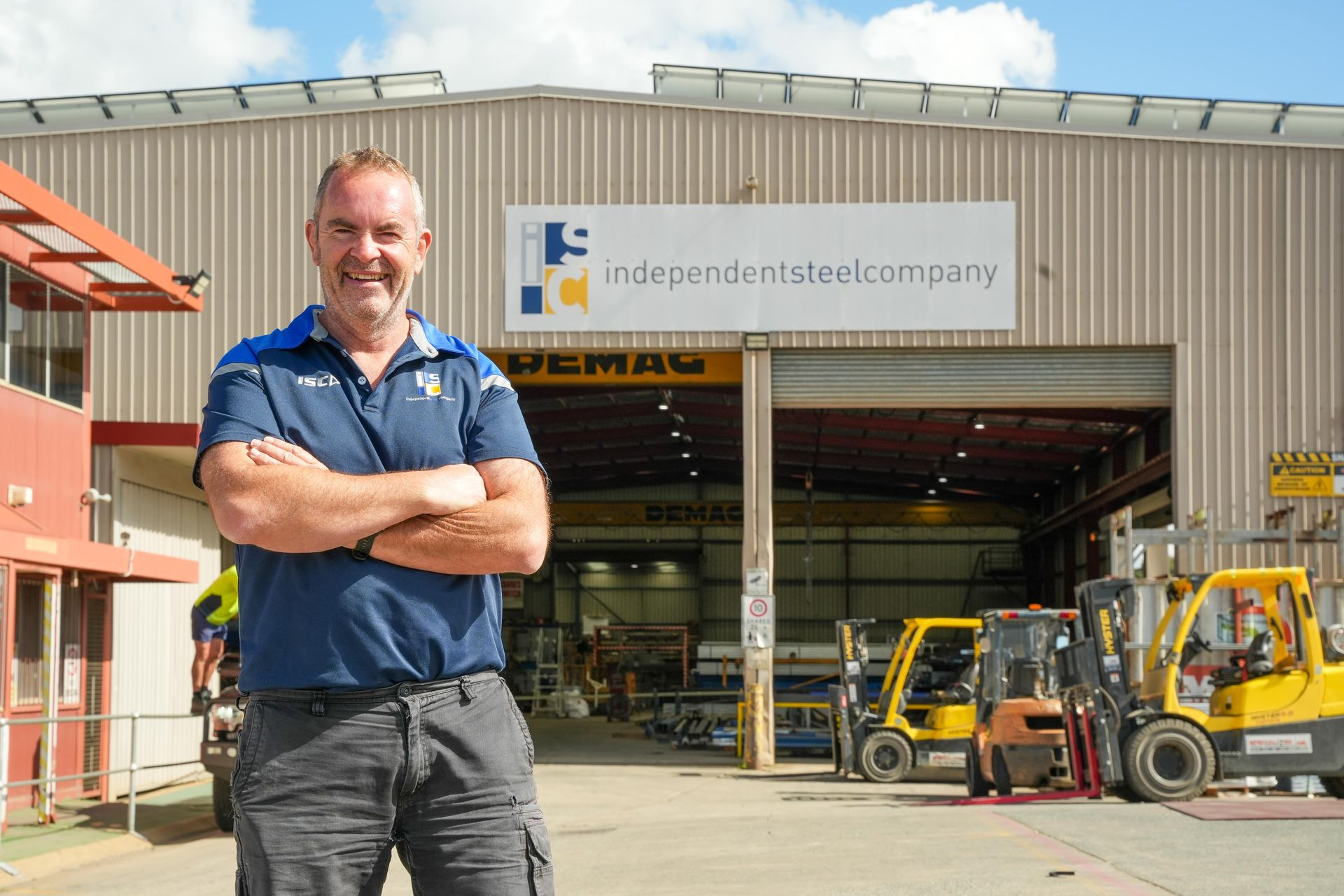 A smiling person stands with arms crossed in front of a steel company warehouse with yellow forklifts parked inside.