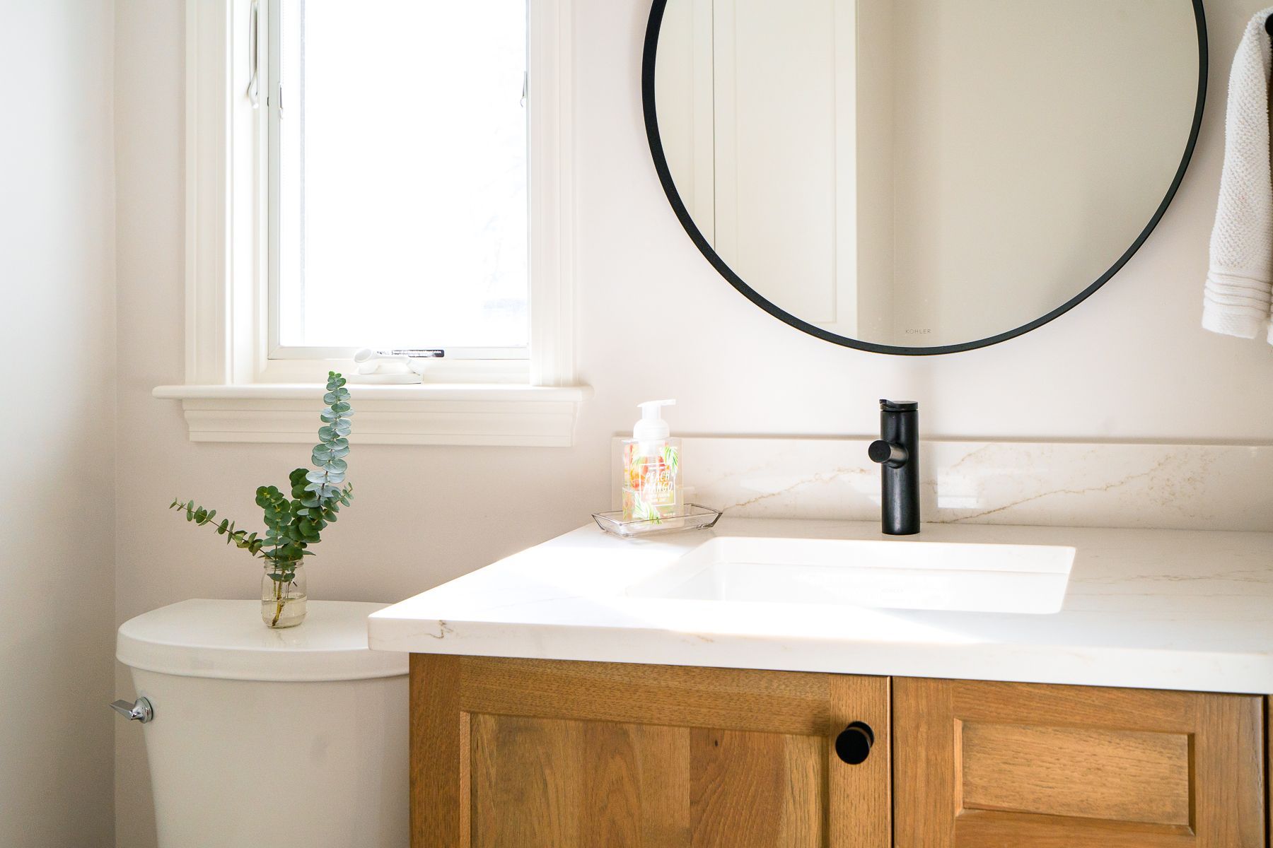 Bright bathroom vanity with round mirror, window, white countertop, and wood cabinet.