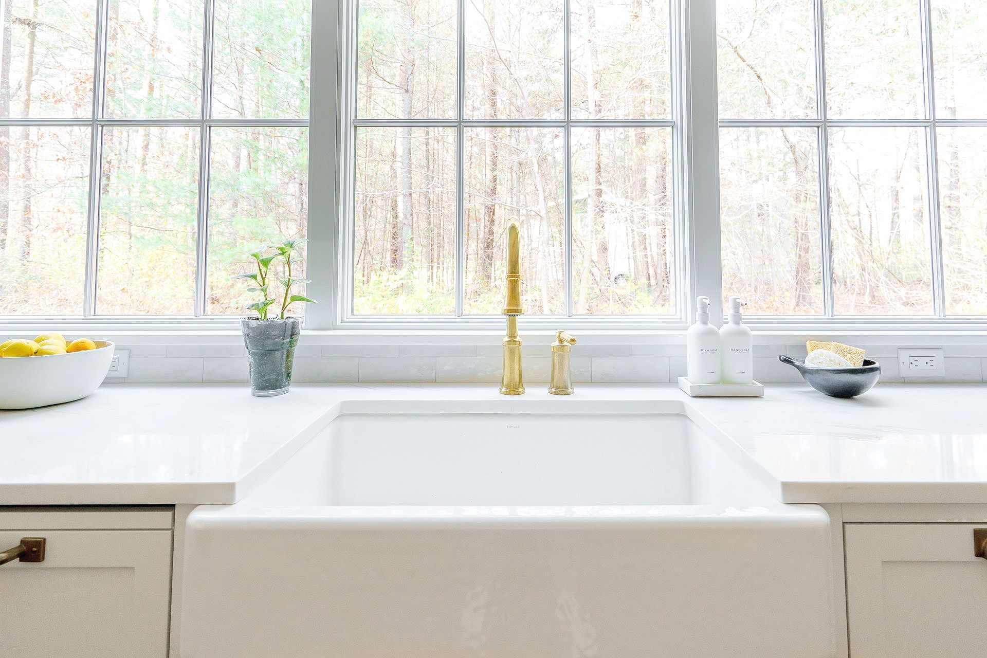 Bright kitchen sink beneath large windows with gold faucet and potted plants on the counter