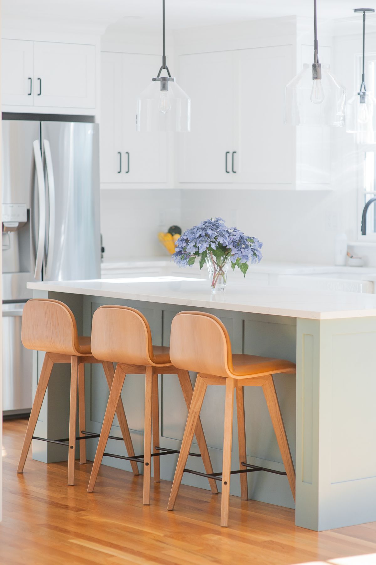 Bright white kitchen with stainless fridge, wooden barstools, and blue flowers on a countertop