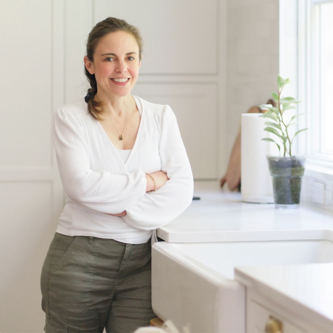 Smiling woman leaning on a white kitchen counter with arms crossed, in a bright room with a potted plant.