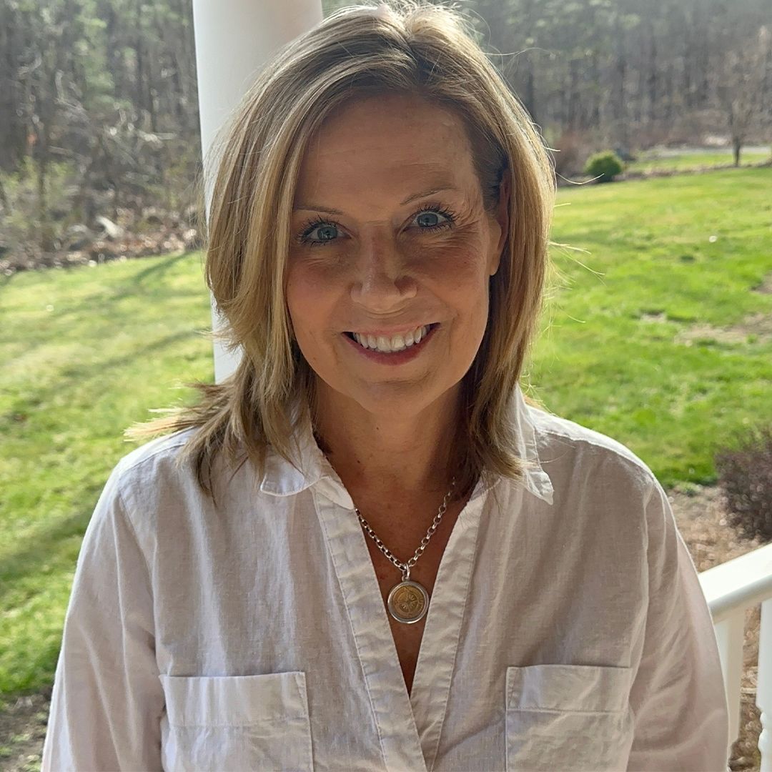 Smiling woman in a white shirt standing on a porch with green lawn and trees behind her