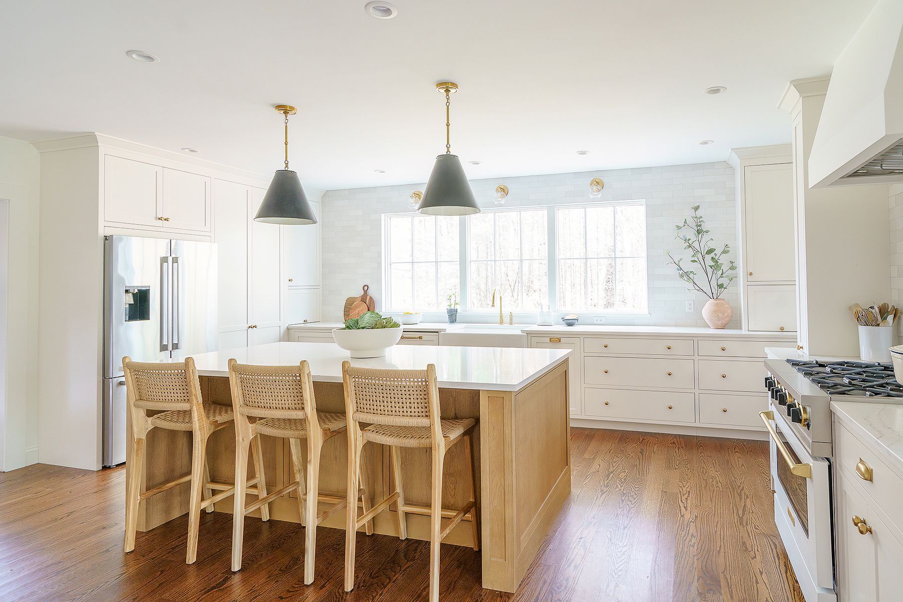 Bright white kitchen with an island, wicker stools, black pendant lights, and sunlit windows