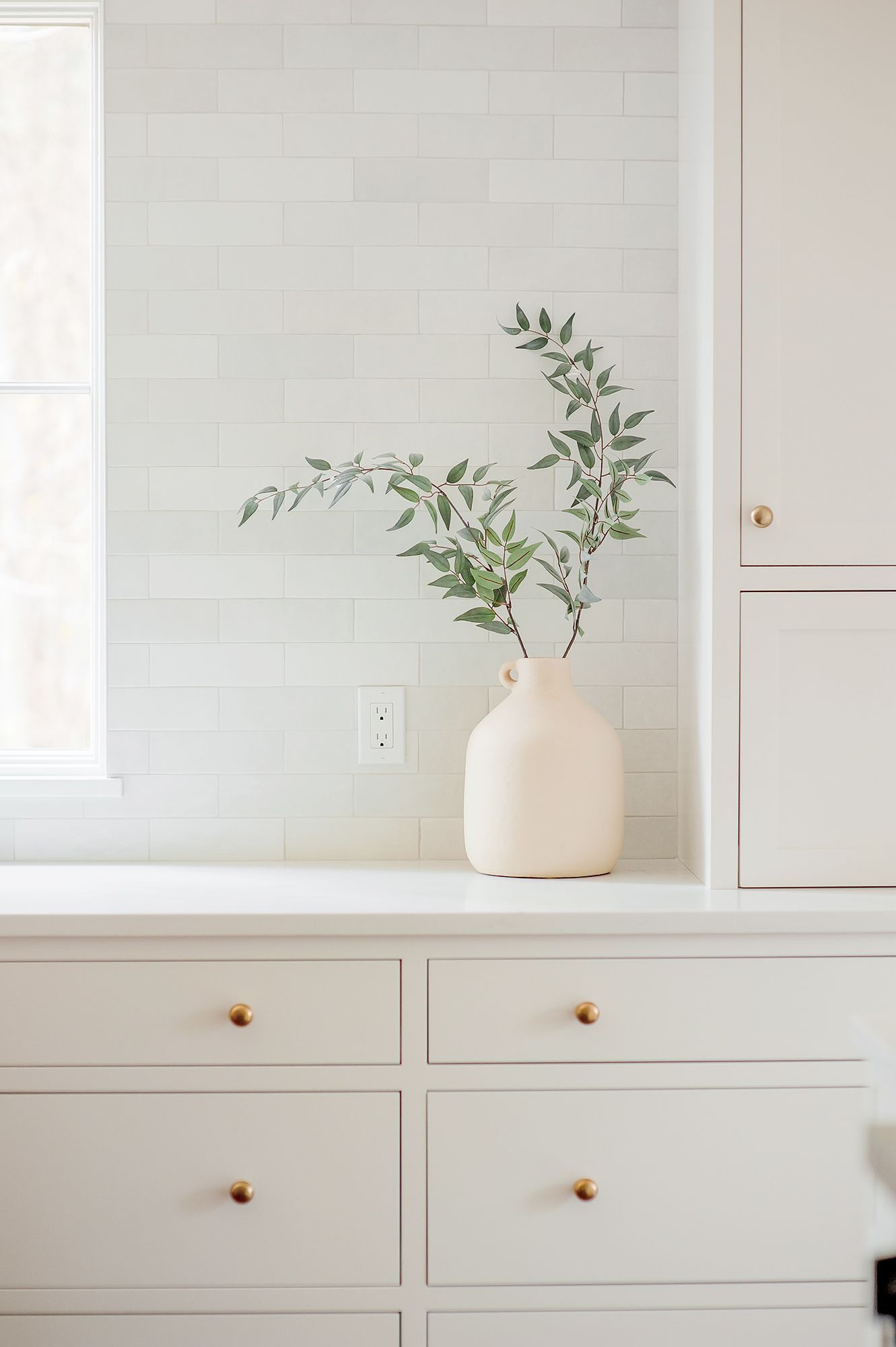 White vase with eucalyptus on a bright kitchen counter, in front of a tiled backsplash and cabinets