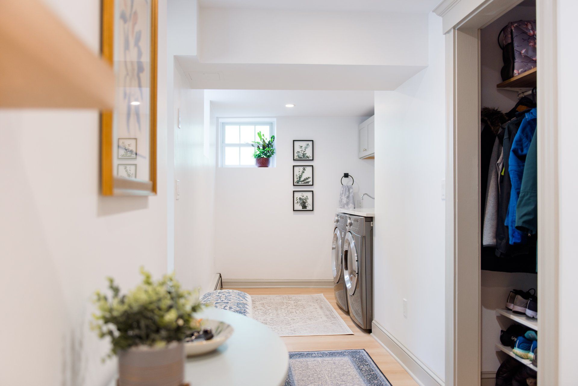 Bright hallway with framed wall art, a rug, potted plants, and an open closet with hanging clothes.