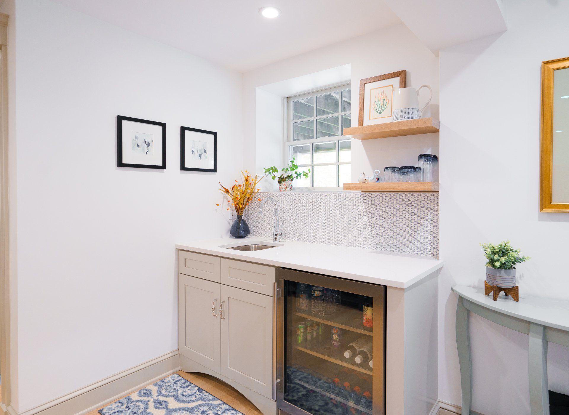 Bright white kitchenette with patterned backsplash, small window, framed art, and a glass-front mini fridge.