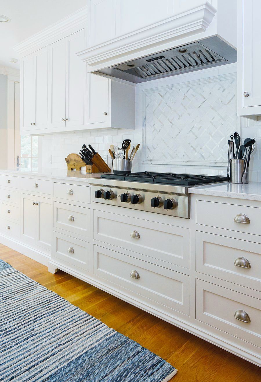 Bright white kitchen with a stainless range, marble backsplash, and blue rug on hardwood floor