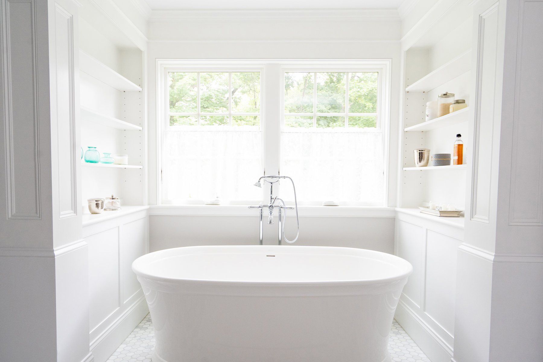 Bright white bathroom with a freestanding tub beneath two windows and built-in shelves.