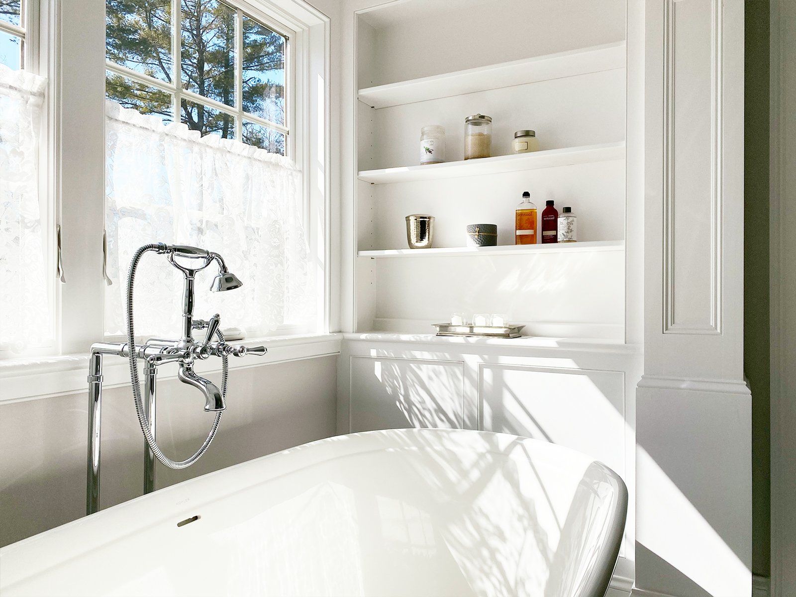 Bright white bathroom with a clawfoot tub, chrome faucet, and built-in shelves by a sunlit window