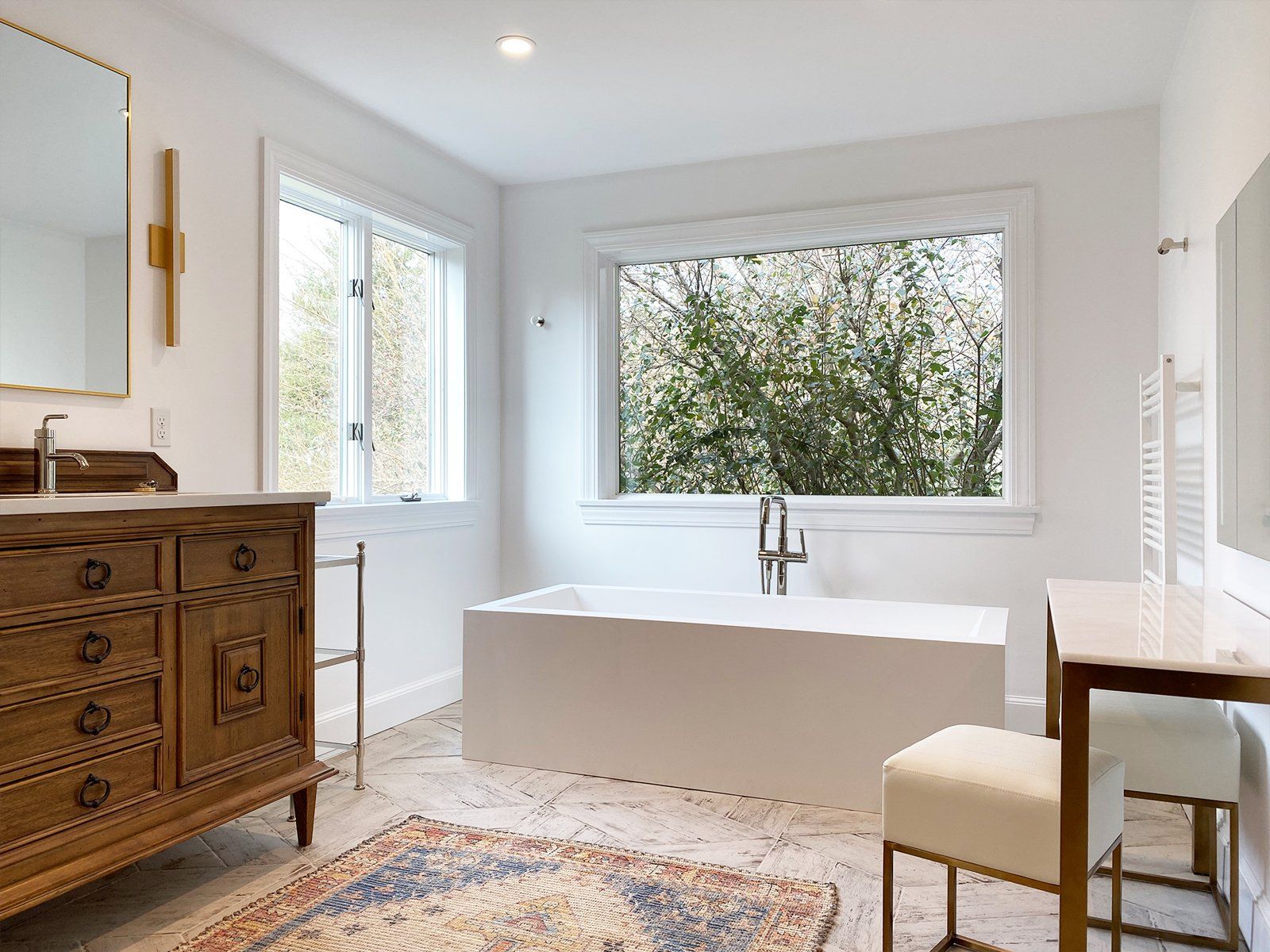 Bright white bathroom with freestanding tub, wood dresser, and large window overlooking greenery