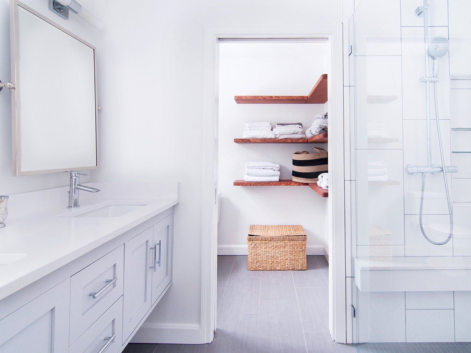 Bright white bathroom with double vanity, shower, and open closet shelves with towels and baskets