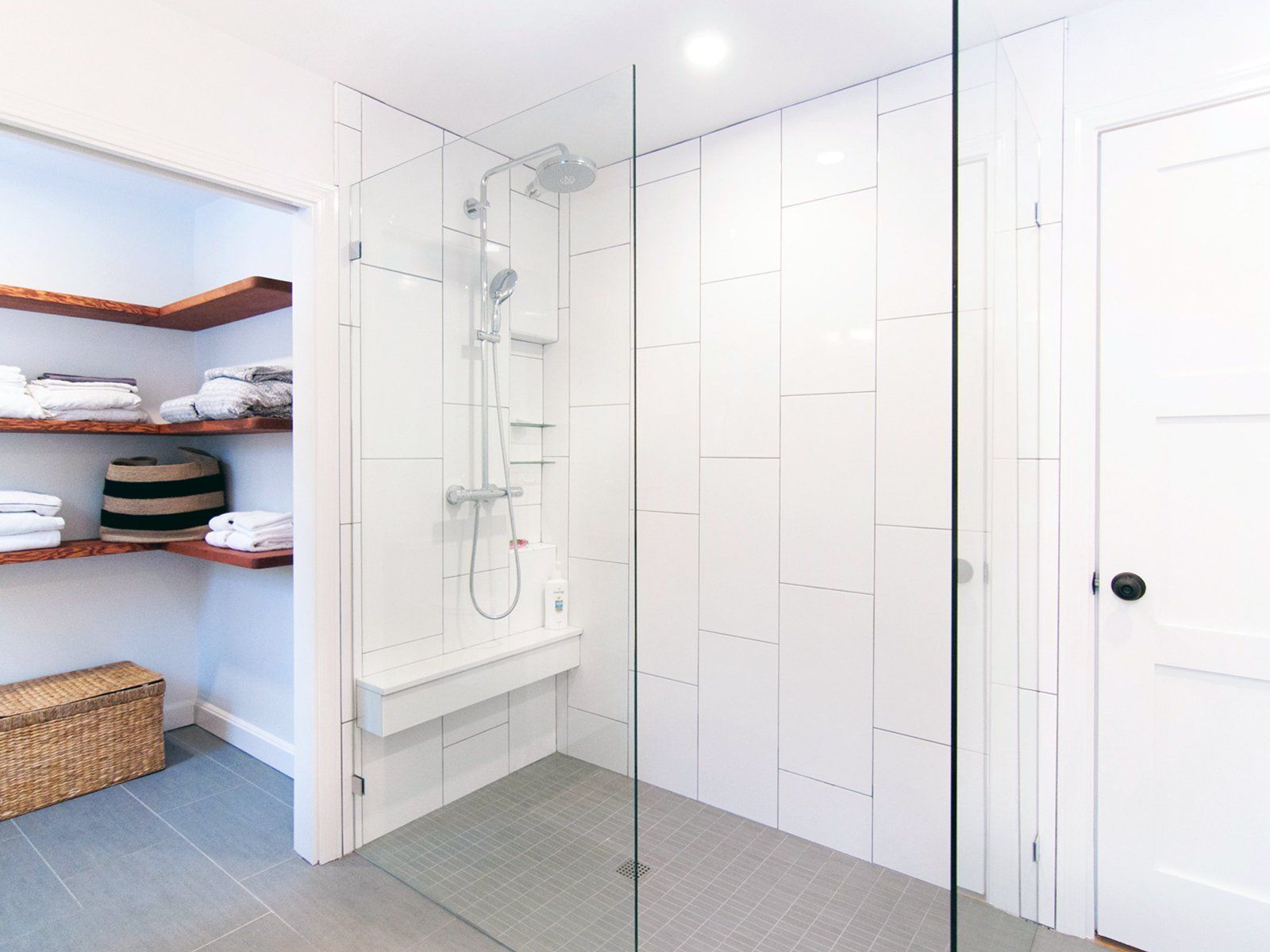 Modern white bathroom with glass shower, gray tile floor, and built-in shelving with towels and baskets