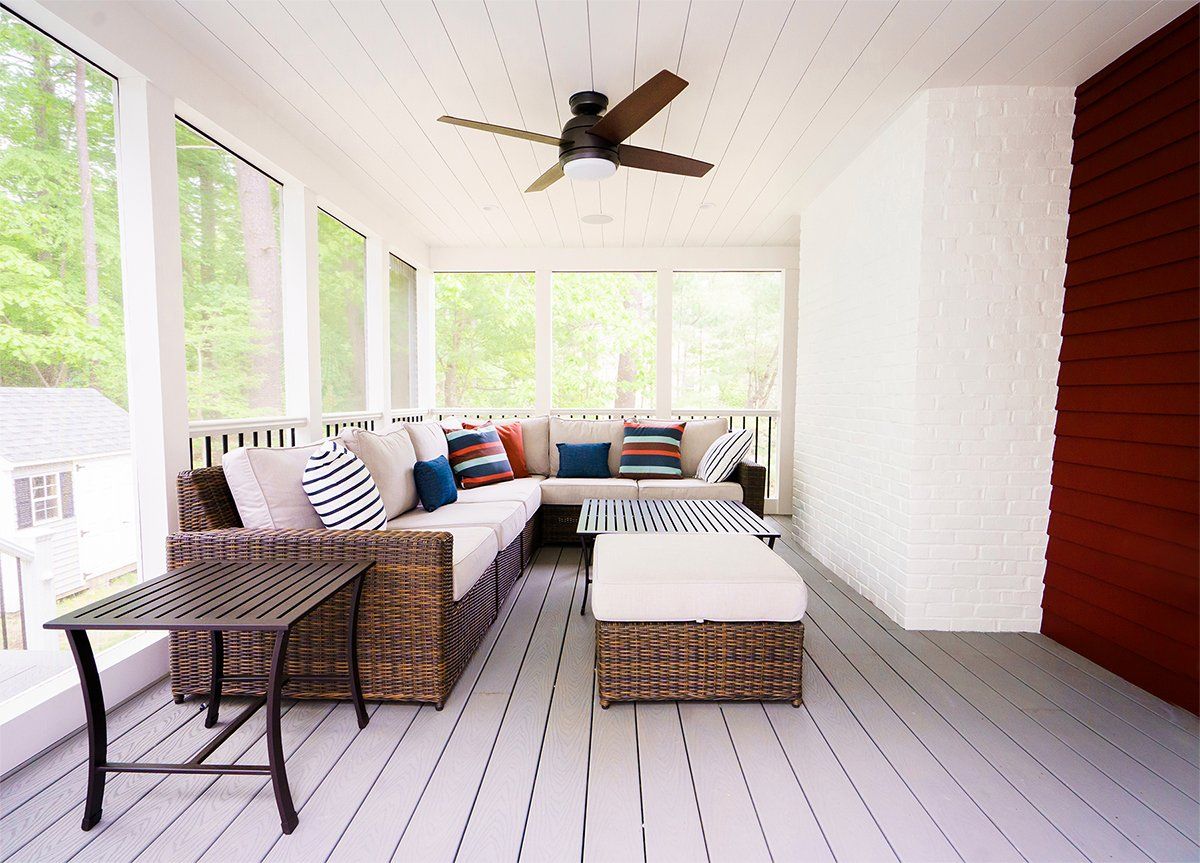 Covered porch with wicker seating, white floorboards, ceiling fan, and bright windows overlooking greenery