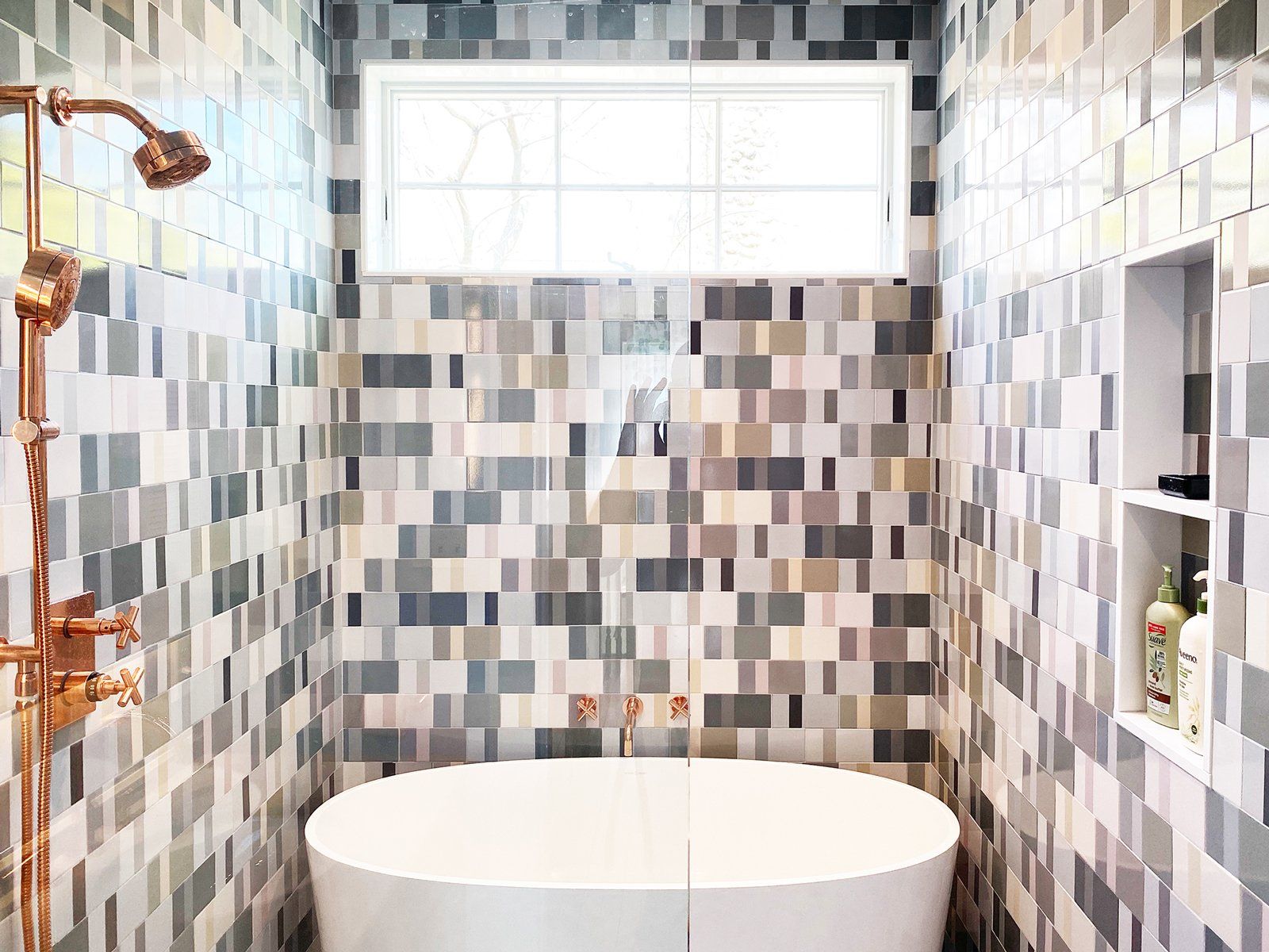 Modern tiled bathroom with a freestanding tub, copper shower fixtures, and a frosted window above.