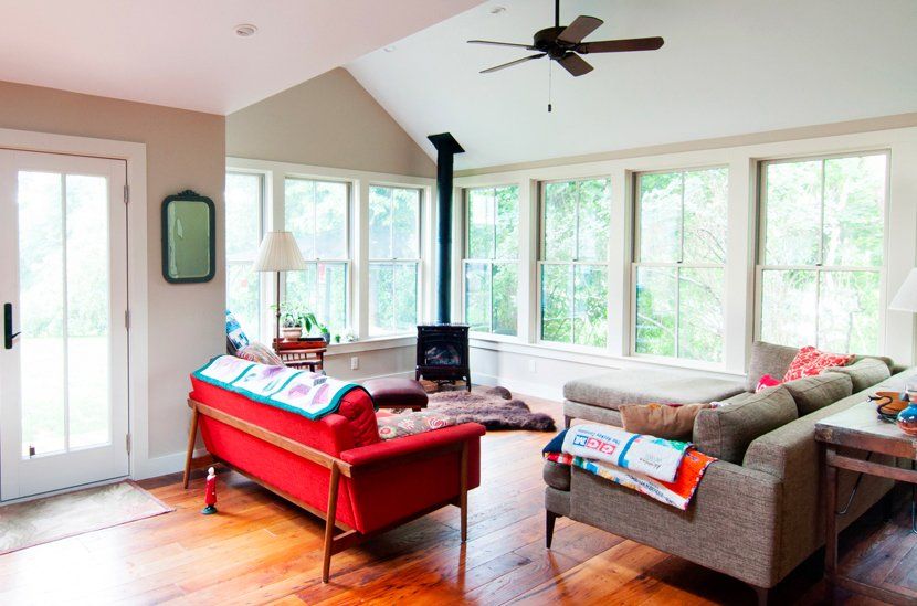 Bright living room with red sofa, gray sectional, wood stove, and large windows overlooking greenery
